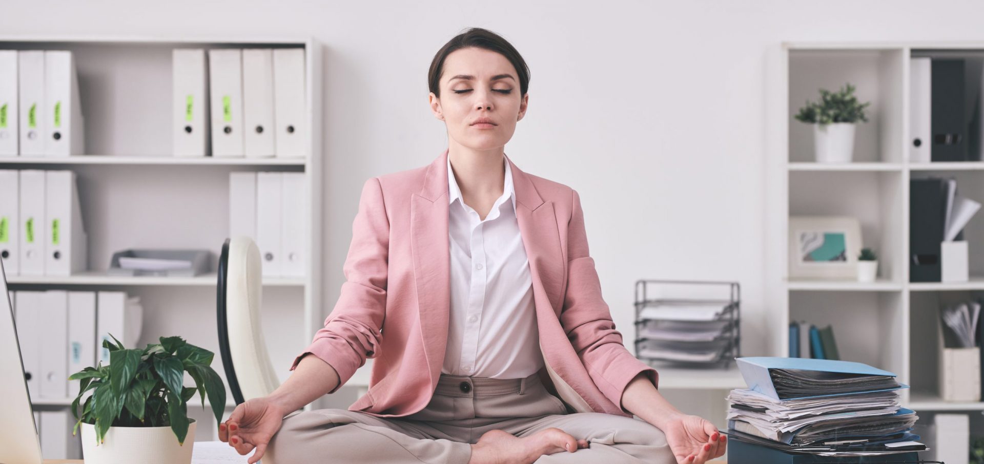 Young spiritual woman in pink jacket sitting in lotus position on table and meditating in office